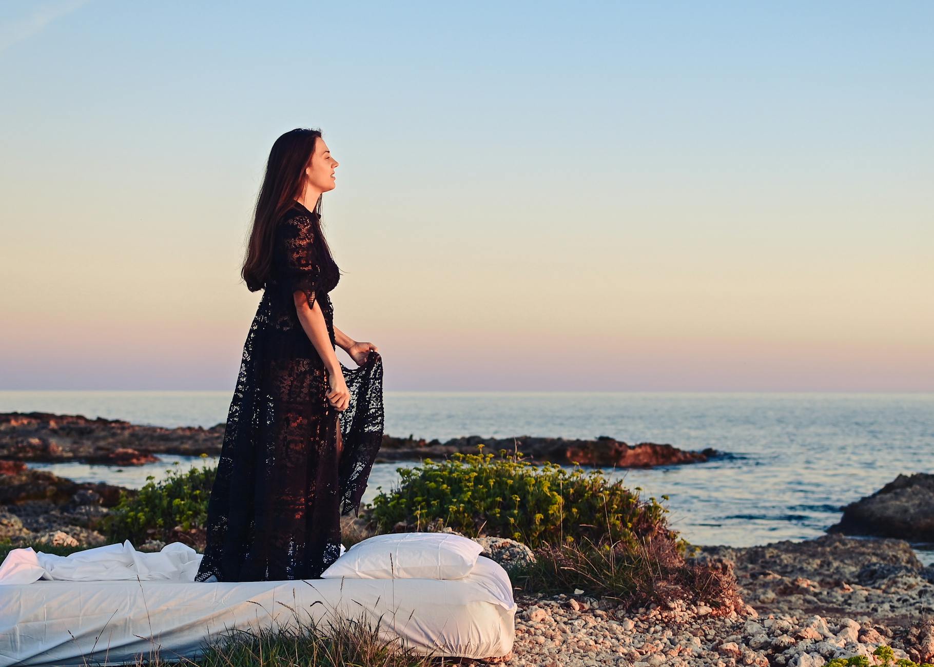 Kobieta w czarnej sukni na skalistym brzegu morza o zachodzie słońca