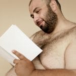 Ranking materacy twardych dla osób 100+ kg: stabilność, wentylacja i trwałość w praktyce Uśmiechnięty brodaty mężczyzna bez koszulki czyta książkę w neutralnym tle
