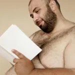 Uśmiechnięty brodaty mężczyzna bez koszulki czyta książkę w neutralnym tle